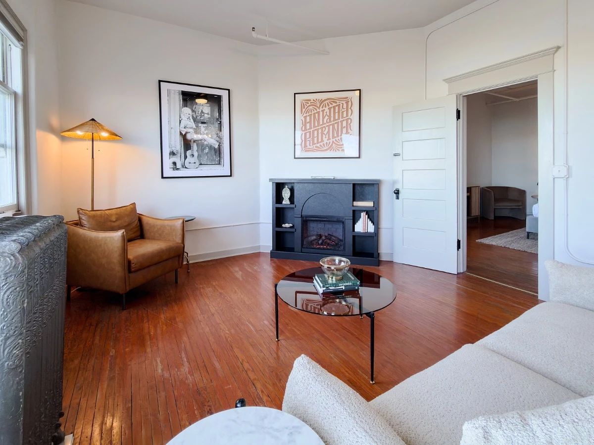 Living room in Room 231 at Hot Lake Springs Resort looking toward electric fireplace, sitting chair to left of fireplace, round coffee table in center, partial sofa on right foreground.