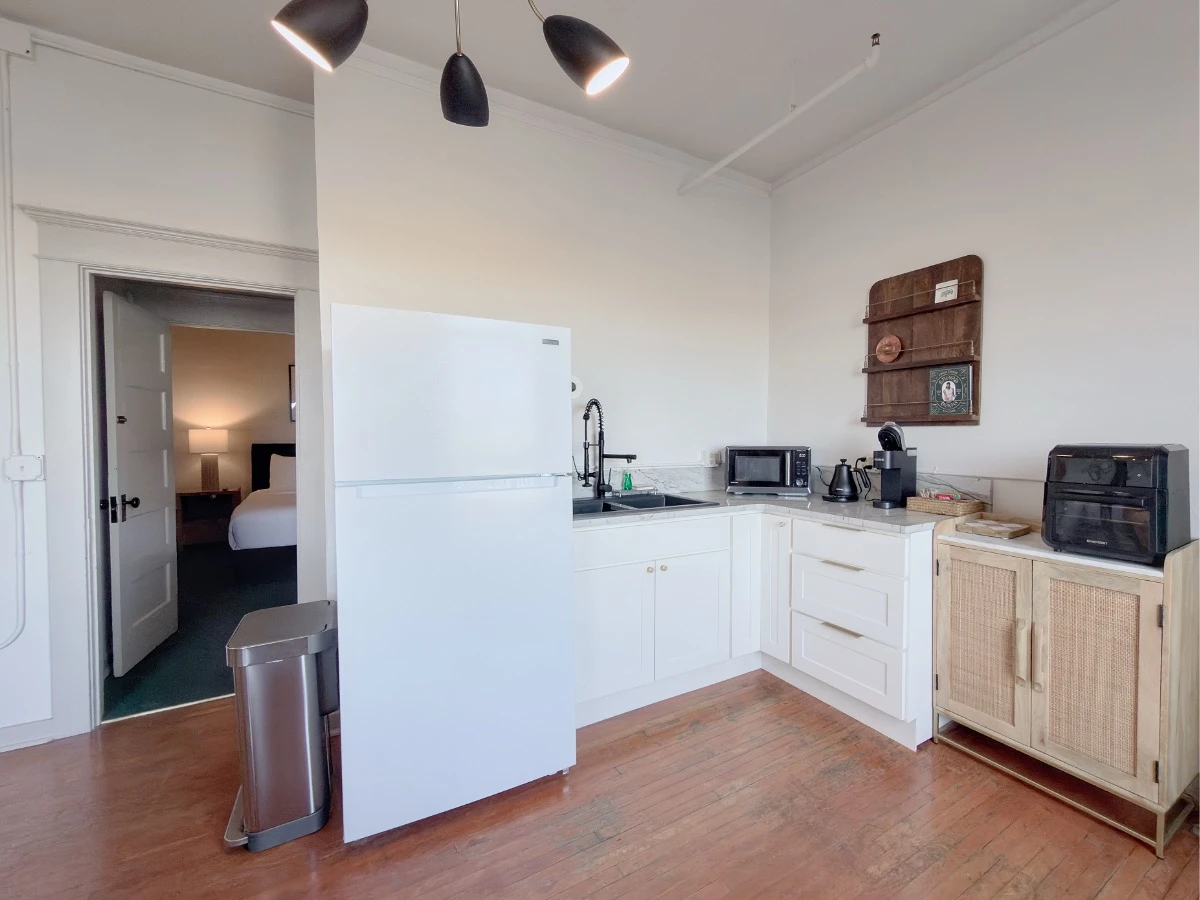 Kitchen in Room 231 at Hot Lake Springs Resort looking toward second room, open door shows partial view of bed. Kitchen has fridge, sink, microwave, electric kettle, coffee machine, and toaster oven on counters.