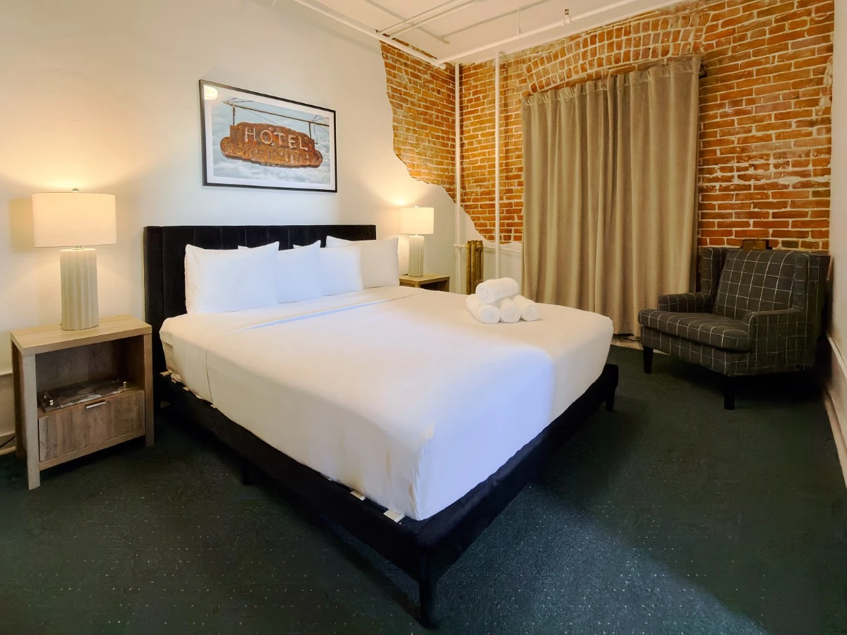 Second bedroom in Room 231 at Hot Lake Springs Resort looking at bed, nightstands with lit lamps on each side, red brick wall on right wall, sitting chair in corner on right side of image.