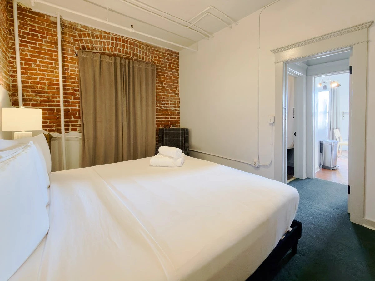Second bedroom in Room 231 at Hot Lake Springs Resort looking across bed out of room, red brick wall on far wall, dark blue/green carpet, window with curtains drawn, sitting chair in background corner.