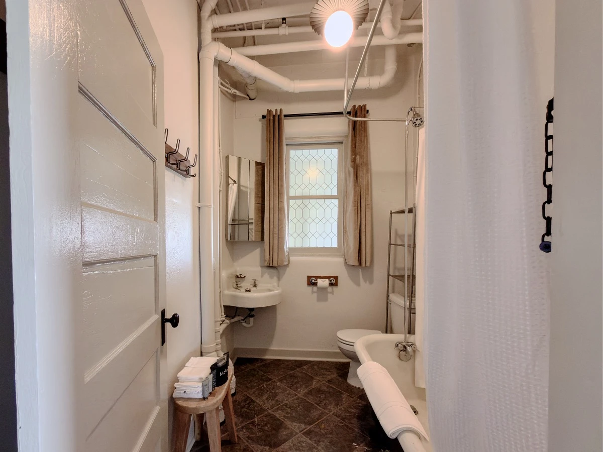 Bathroom in Room 231 at Hot Lake Springs Resort showing corner sink, toilet, and clawfoot soaking tub/shower combo, mirror above sink, window on far wall by sink and toilet.