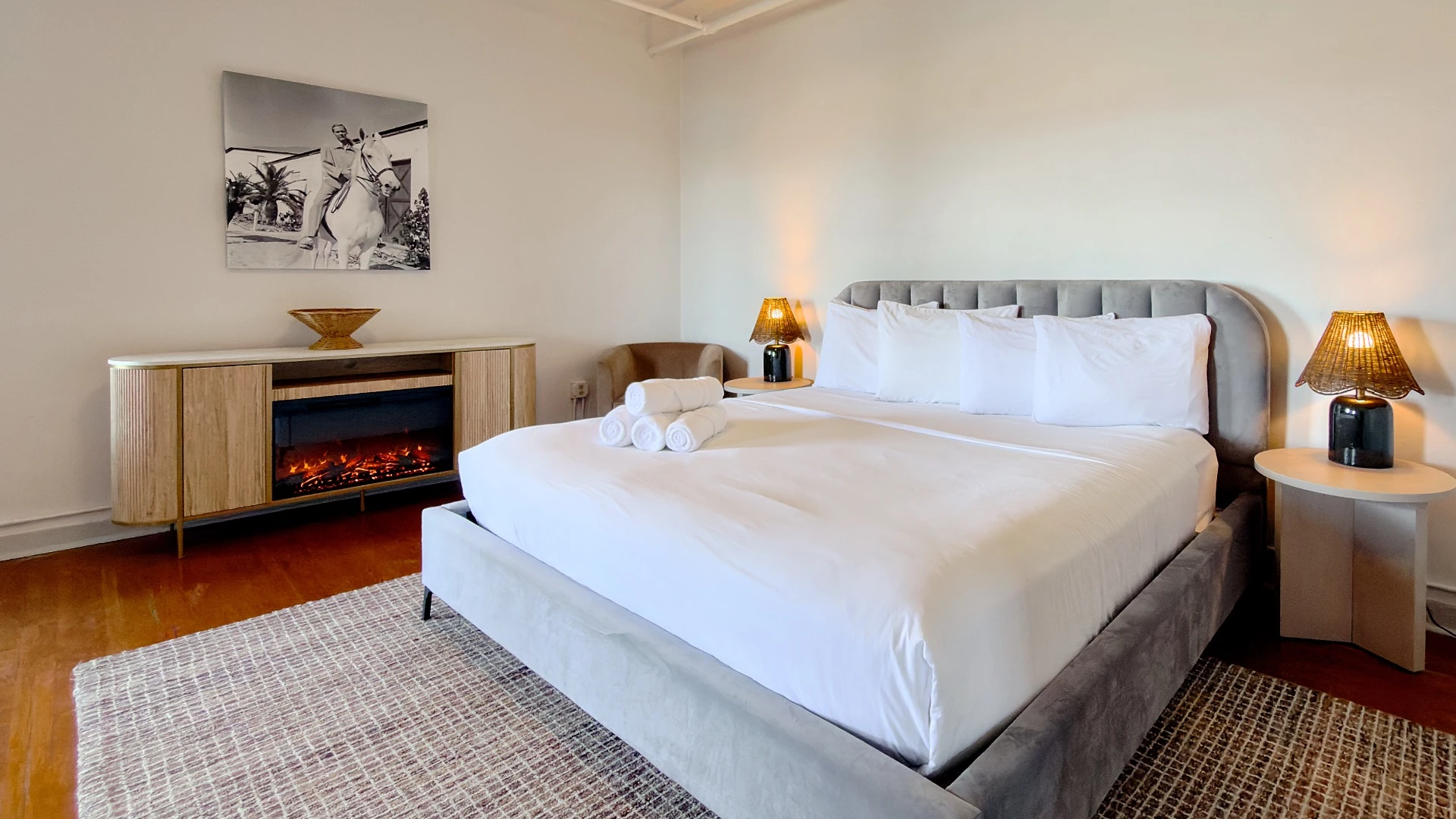 Master bedroom in Room 231 at Hot Lake Springs Resort with electric fireplace next to bed, black and white photo of man on horse above fireplace, wood floors with grey rug under foot of bed, sitting chair in back corner, nightstands with lit lamps on each side.