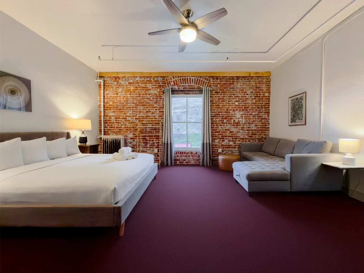 View toward window on red brick wall in Room 324 at Hot Lake Springs Resort, bed on left, grey sofa with chaise lounge on right, end table with lit lamp next to sofa.