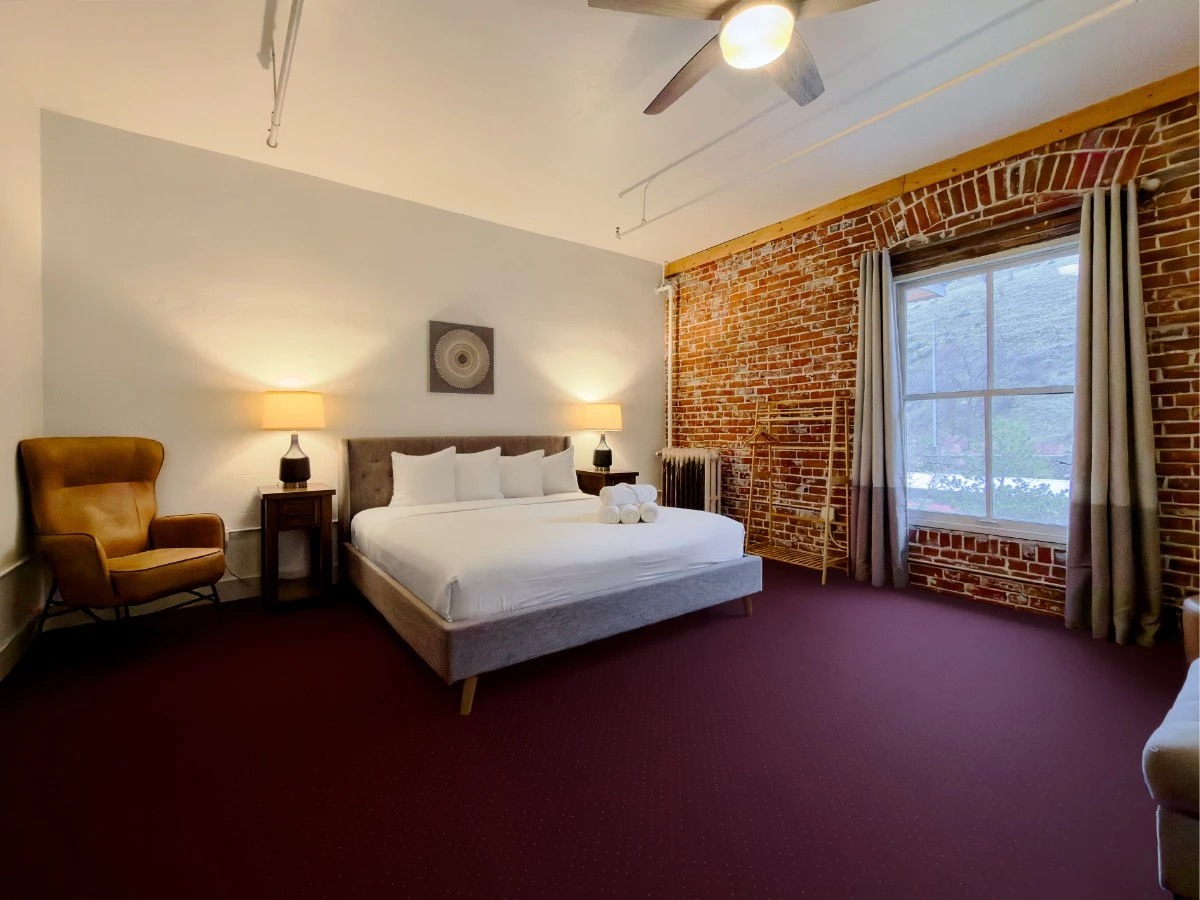 View from corner shower in Room 324 at Hot Lake Springs Resort toward bed and window, high-back sitting chair to left of bed, red brick wall with window on right of bed.