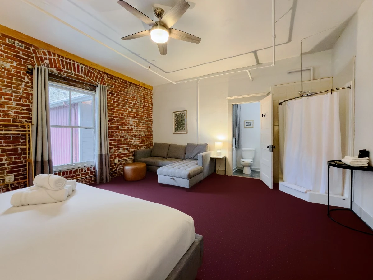 View toward couch in Room 324 at Hot Lake Springs Resort with open bathroom door to right of couch showing toilet visible, red exposed brick wall with window on left, corner shower in room on right, partial foot of bed in left foreground.
