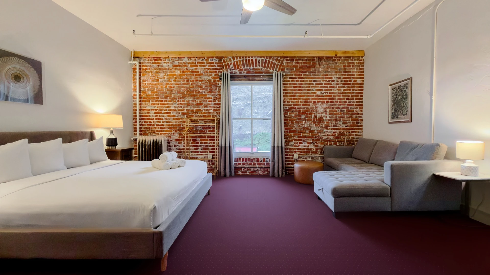View toward window on red brick wall in Room 324 at Hot Lake Springs Resort, bed on left, grey sofa with chaise lounge on right, end table with lit lamp next to sofa.