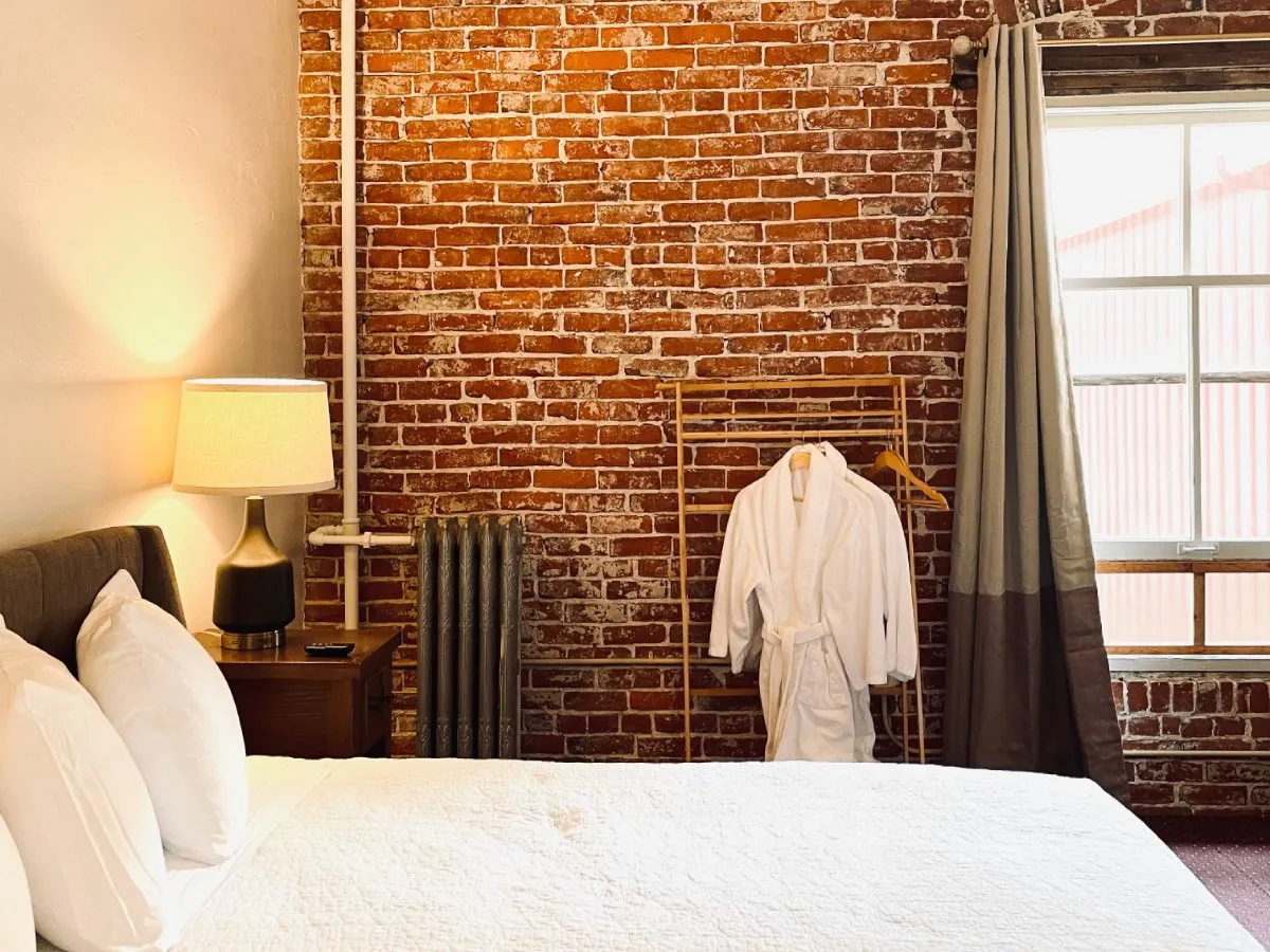 Interior of Room 324 at Hot Lake Springs Resort, bed with white linens, bedside lamp lit, unique brick wall, and white bathrobe hanging.