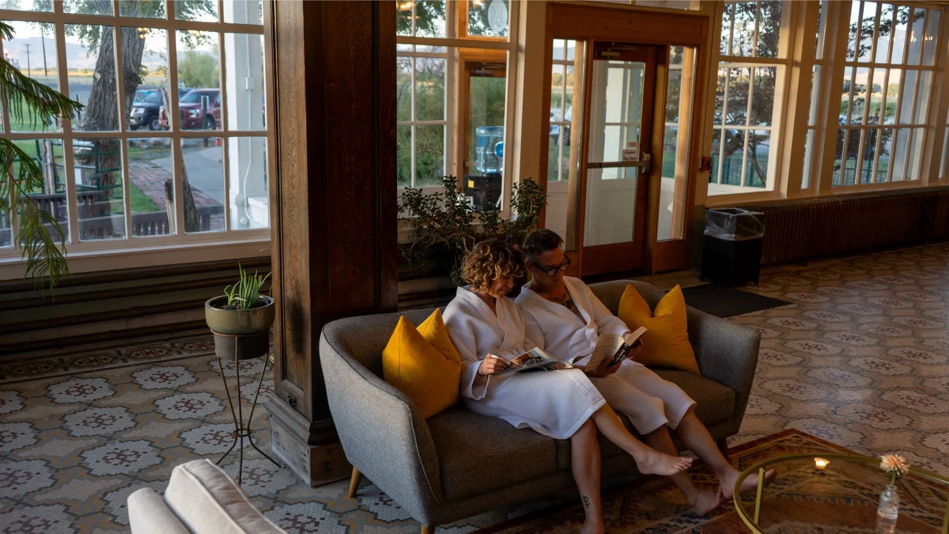 Couple relaxing in white bathrobes on sofa in main gallery lounge at Hot Lake Springs Resort, reading by tall windows with historic tile floors, cool after-sunset lighting.