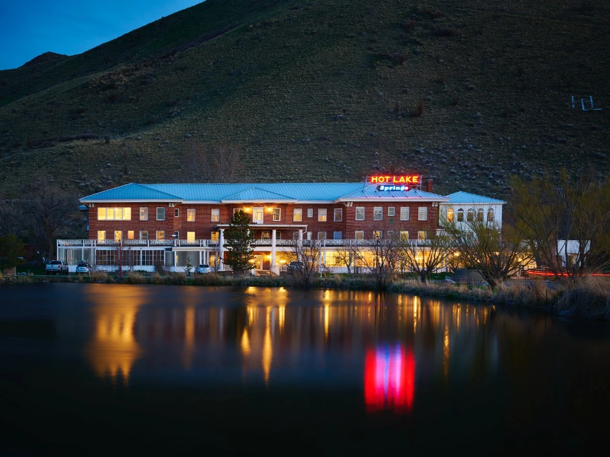 Front exterior of Hot Lake Springs Resort at dusk, red brick facade with iconic neon sign lit up and soft reflection in Hot Lake.