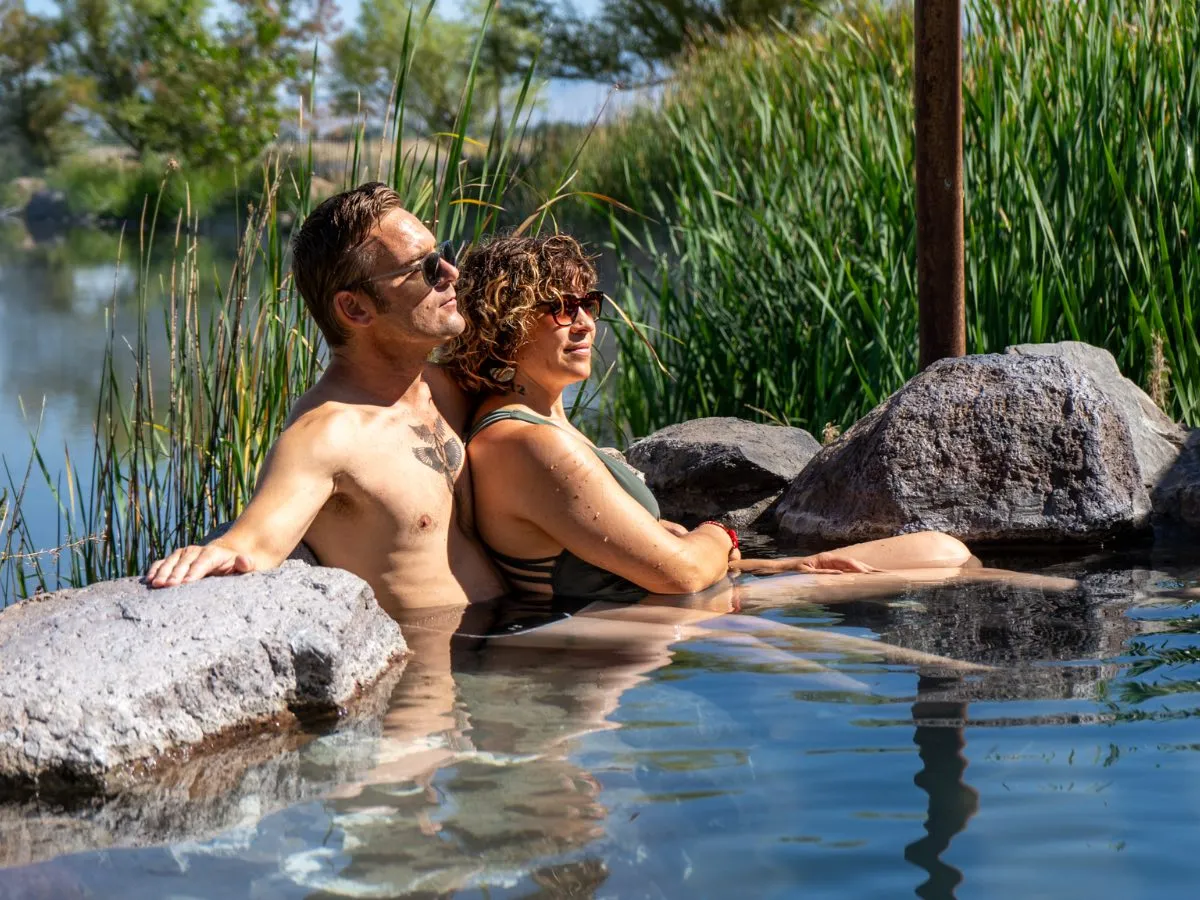 Couple wearing sunglasses soaking in rock-style tub at Hot Lake Springs Resort, summer greenery in background, clear water with reflections.