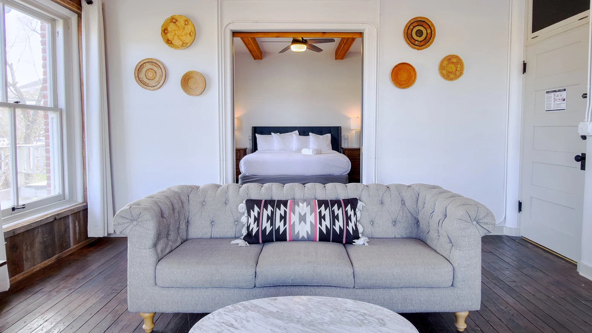 Well-lit historic lodge room at Hot Lake Springs Resort with grey sofa, marble coffee table, dark wood floors, rustic timber beams, and bed visible through open sliding doors.