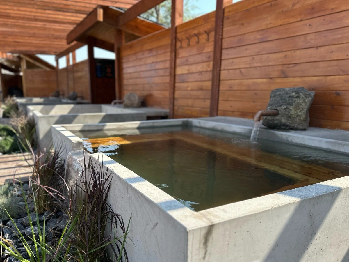 Angled view of square soaking tub at Hot Lake Springs Resort, plant accents at spill-over, matching tubs behind, wood fence on right, pergola overhead.