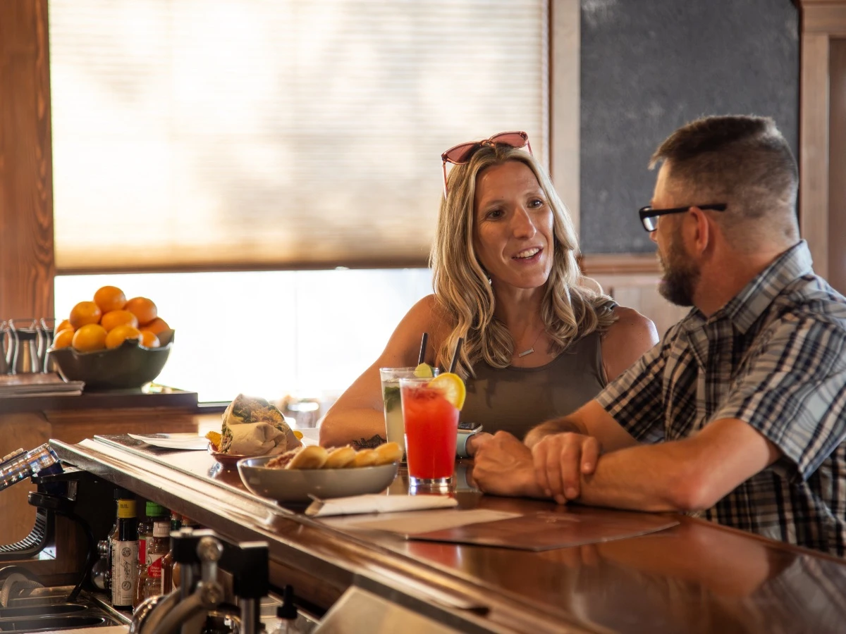 Couple enjoying drinks and food at the bar top in Thermal Pub & Eatery at Hot Lake Springs Resort, glowing window with drawn blind in background.