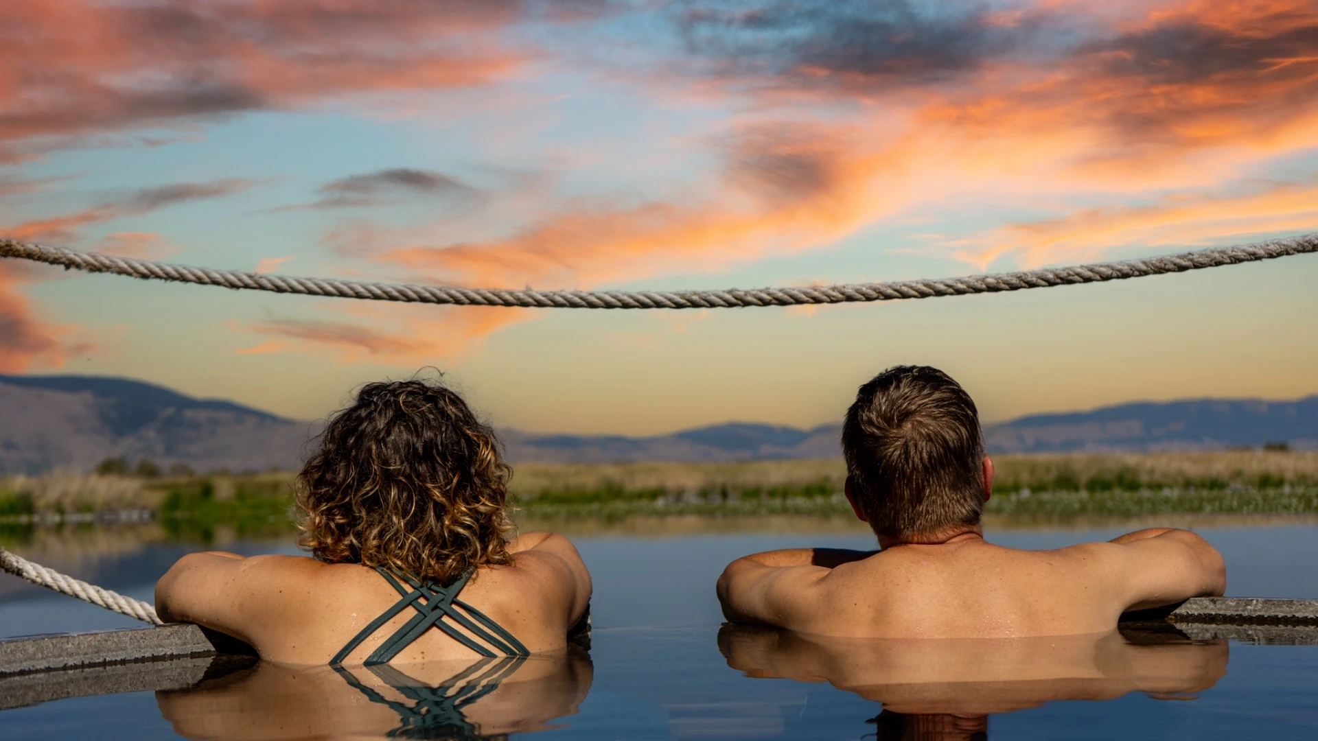 Couple soaking in round tub at Hot Lake Springs Resort, backs to camera, watching sunset over distant mountains with copper clouds and calm lake water.