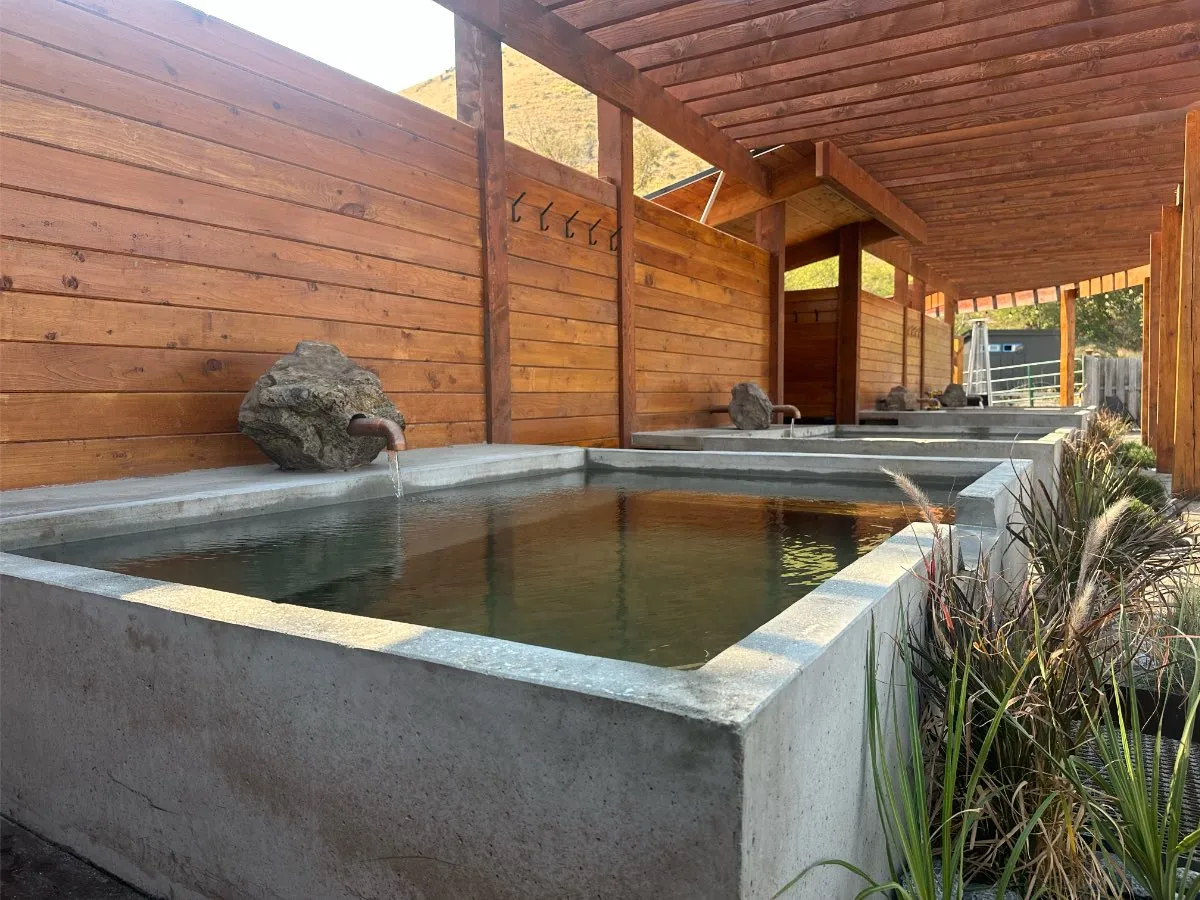 Angled view of square soaking tub at Hot Lake Springs Resort, plant accents at spill-over, matching tubs behind, wood fence on left, pergola overhead.