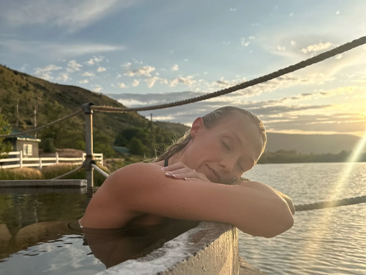 Woman leaning over edge of round soaking tub at Hot Lake Springs Resort, head on arms, summer sunset beginning in background.