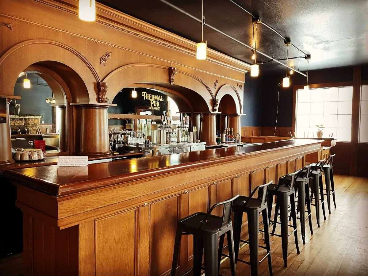 Wood bar top with stools in the Thermal Pub & Eatery at Hot Lake Springs Resort, mirror in background displaying logo, beer taps, and glassware.