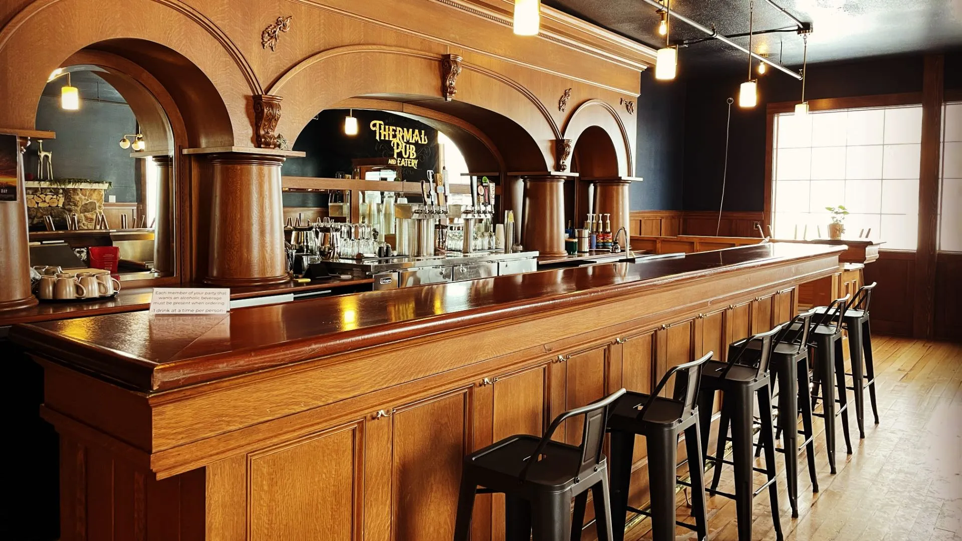 Wood bar top with stools in the Thermal Pub & Eatery at Hot Lake Springs Resort, mirror in background displaying logo, beer taps, and glassware.