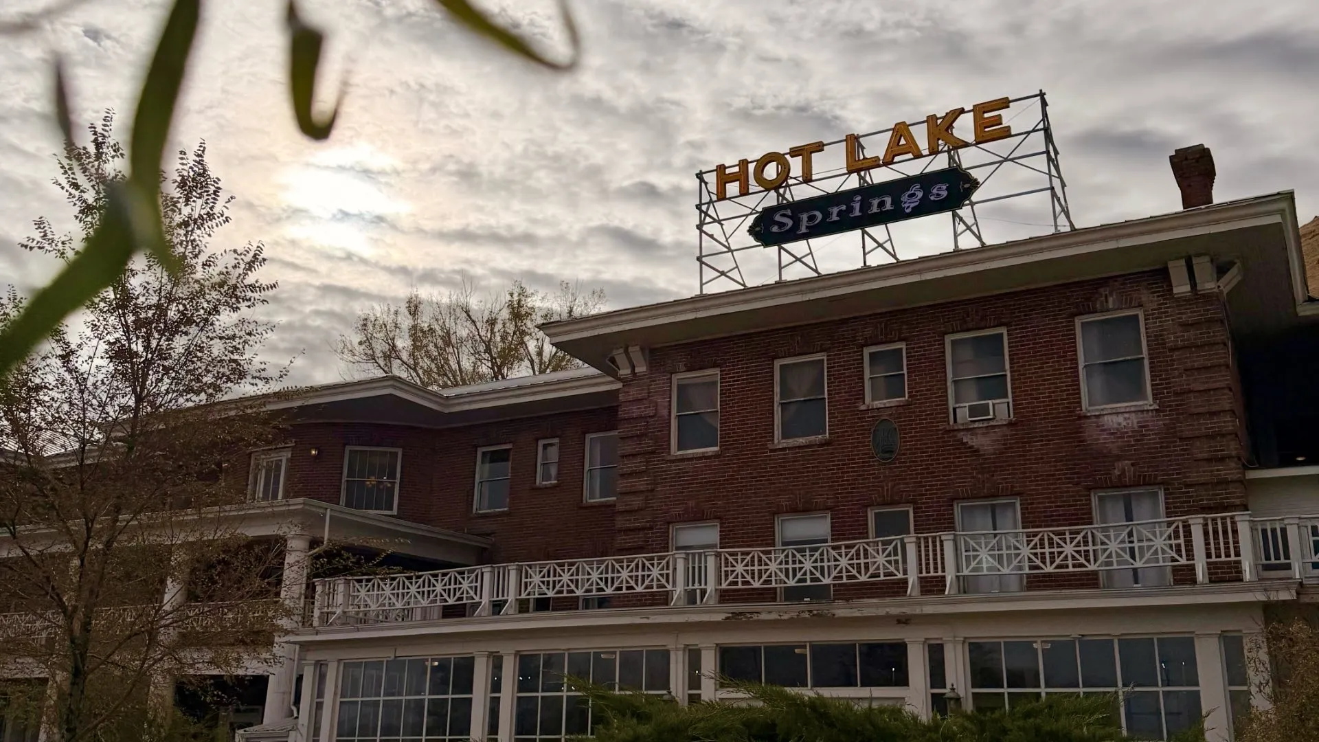 Cloudy daytime view looking up at the front of Hot Lake Springs Resort, historic red brick facade with neon sign on top.