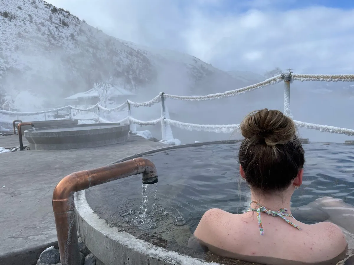View over woman’s shoulder in soaking tub at Hot Lake Springs Resort on frosty day, ice on ropes, steam from Hot Lake, snow on distant mountains.