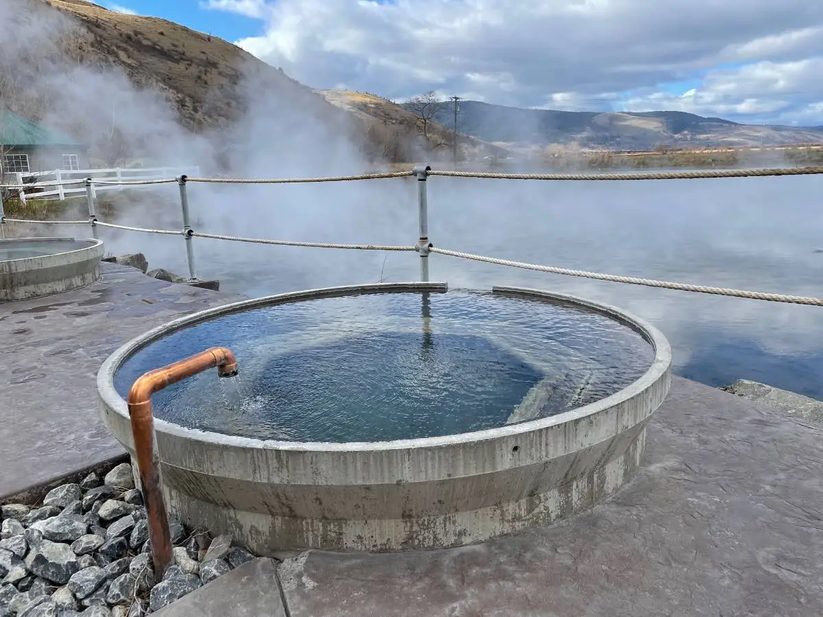 Clear summer water in round soaking tub at Hot Lake Springs Resort, white steam rising from Hot Lake in background.