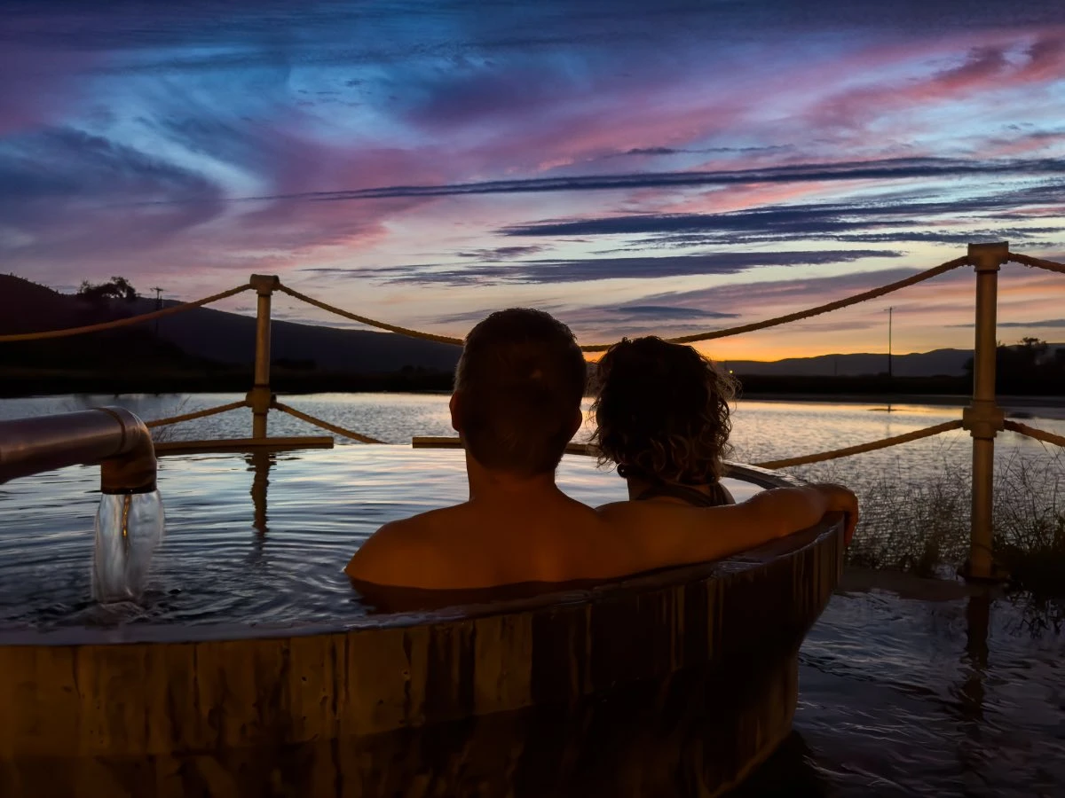 Couple in elevated round soaking pool at Hot Lake Springs Resort at late sunset, man’s arm around woman, backs to camera, purple and gold clouds above.