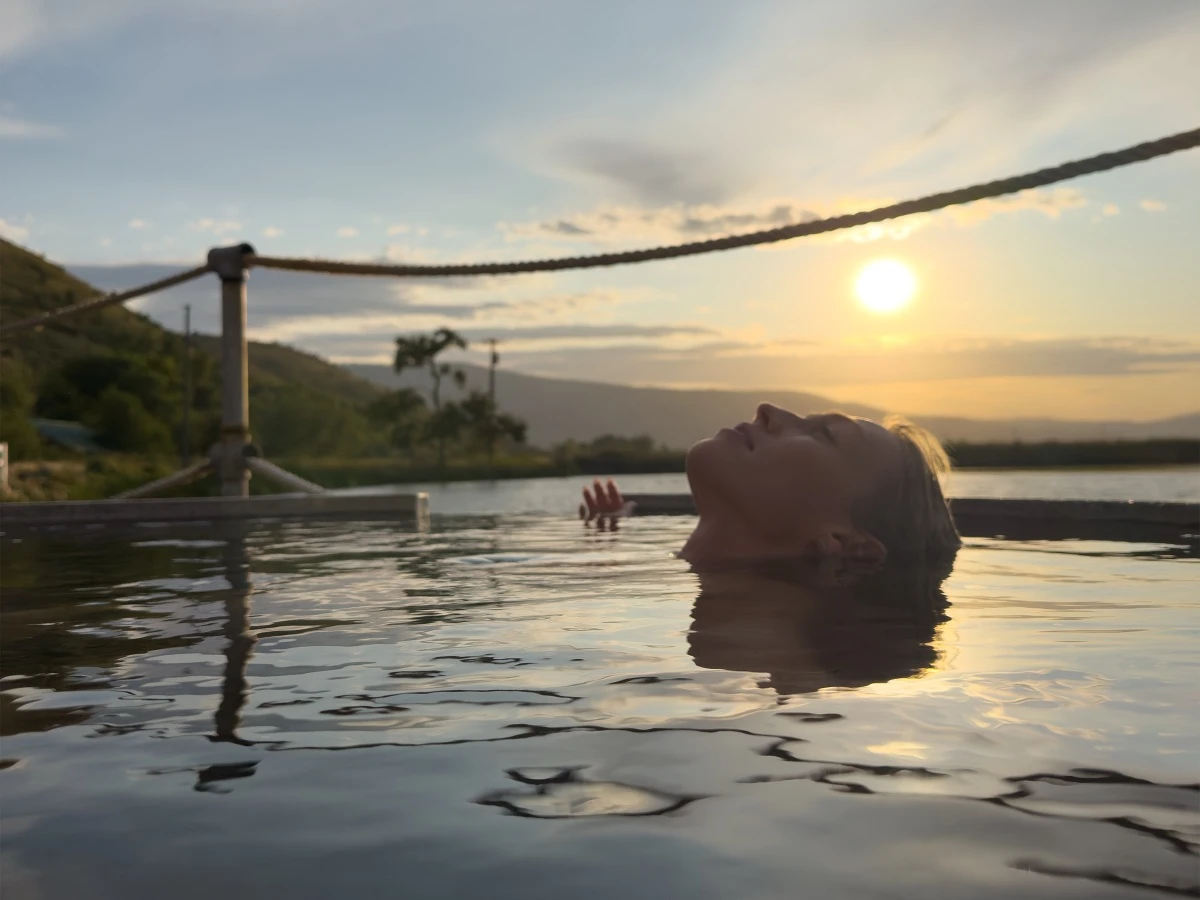 Woman soaking in tub at Hot Lake Springs Resort at late sunset, head visible looking skyward, sun reflecting on water.