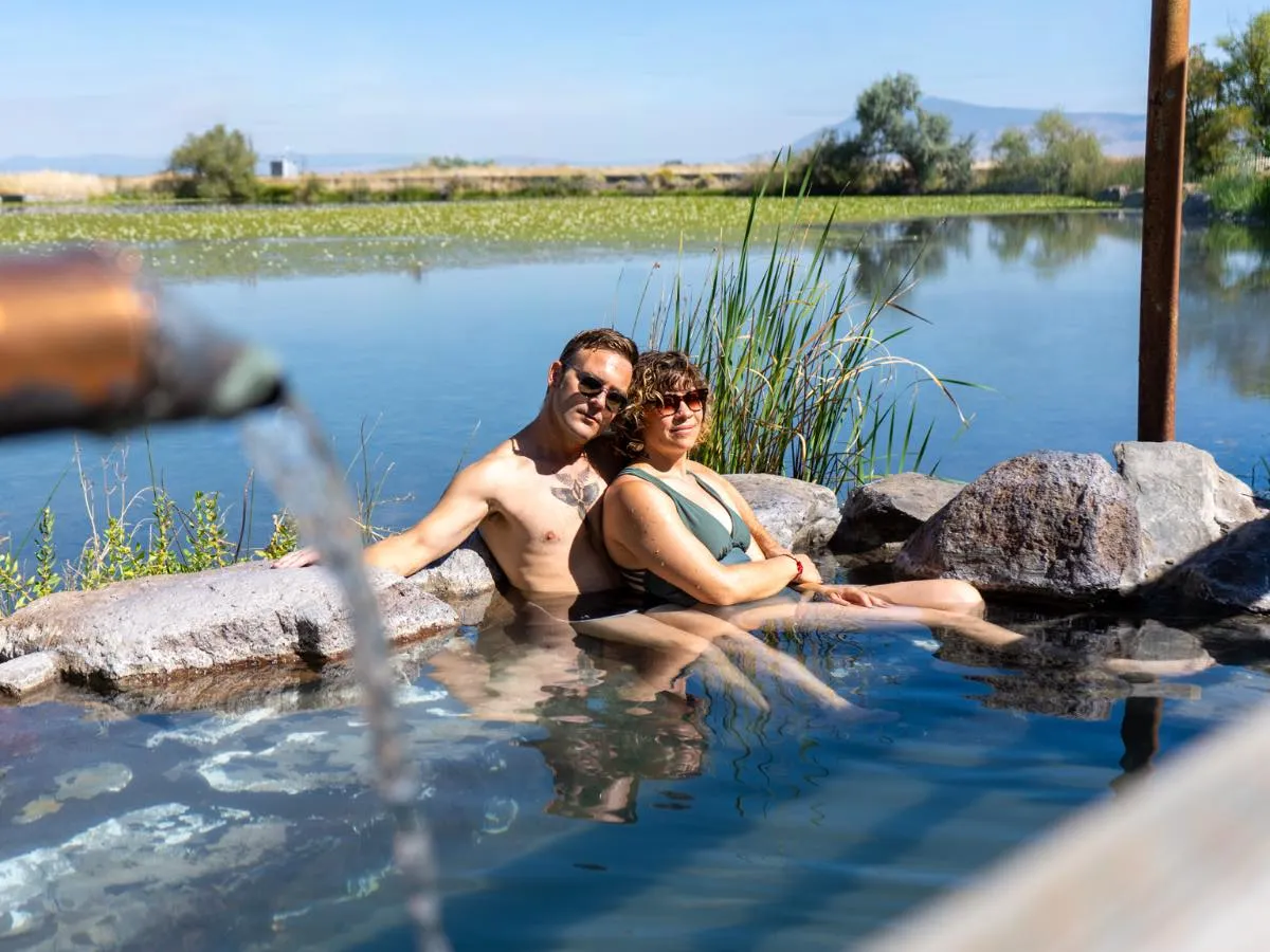 Couple soaking in rock-style tub at Hot Lake Springs Resort, faucet running on left, still Hot Lake and summer greenery in background.