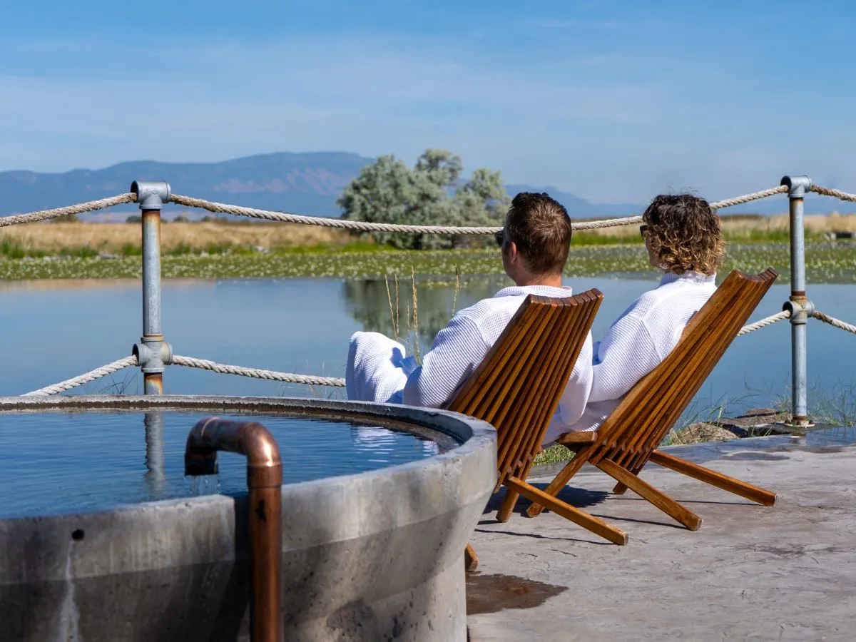 Couple in white robes relaxing in Adirondack chairs beside elevated round soaking pool at Hot Lake Springs Resort, summer day with blue sky and calm water.