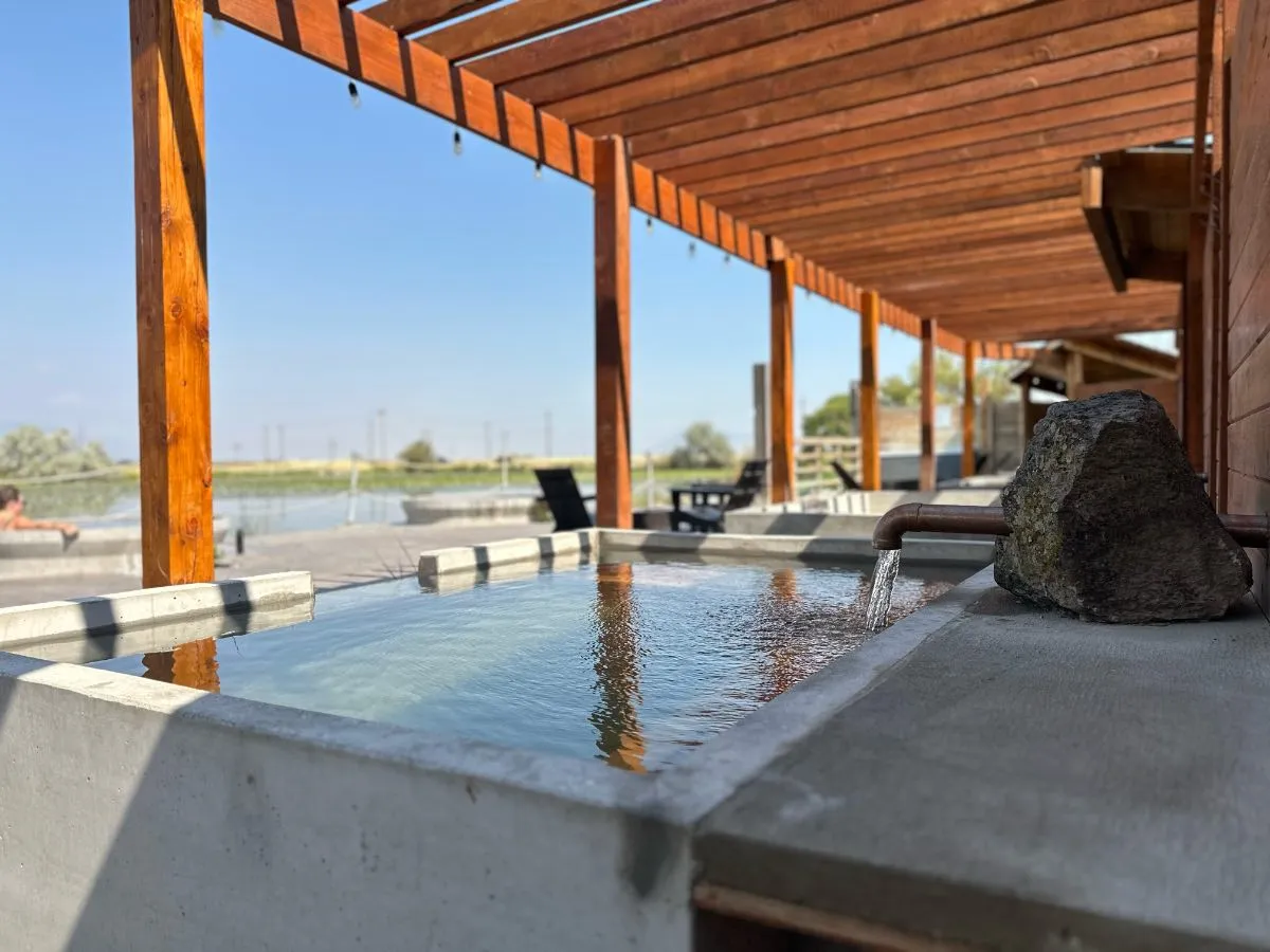 Side view of square soaking tub at Hot Lake Springs Resort with copper faucet coming through stone, wood pergola above and fence on right, matching tubs across, summer valley beyond.