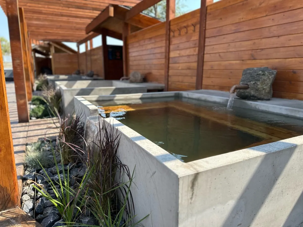 Angled view of square soaking tub at Hot Lake Springs Resort, plant accents at water spill-over, matching tubs behind, wood fence on right, pergola overhead.