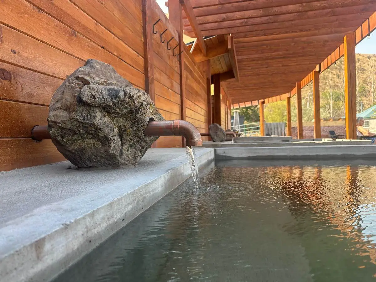 Side view of square soaking tub at Hot Lake Springs Resort with copper faucet coming through stone, wood pergola above and fence on left, matching tubs extending into background.