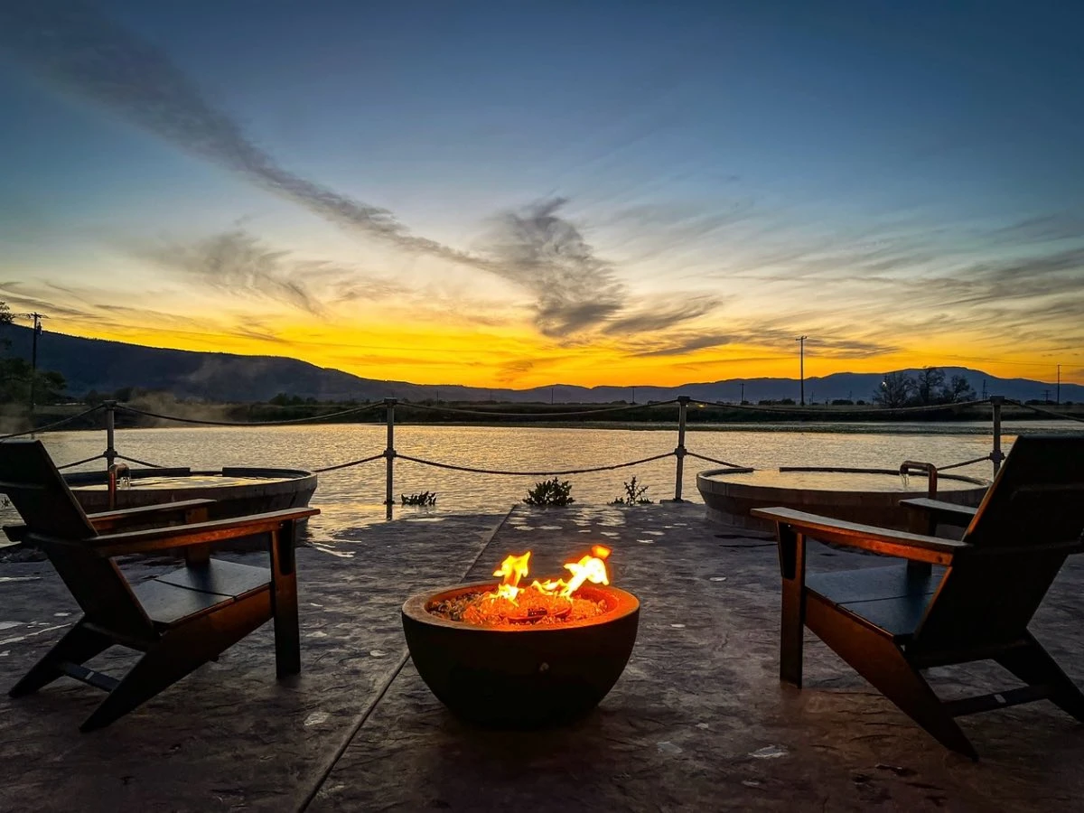 Sunset from soaking area at Hot Lake Springs Resort, Adirondack chairs with fire pit, golden glow on mountains, dark blue sky, and subtle yellow light on Hot Lake.
