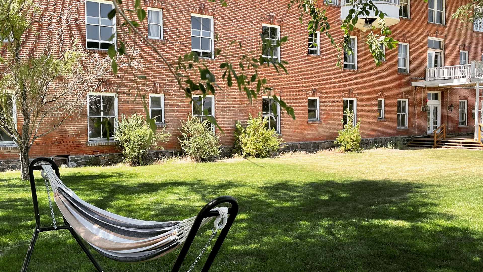 Sunny East lawn at Hot Lake Springs Resort with hammock, green grass, tree shade, and historic red brick lodge in background.