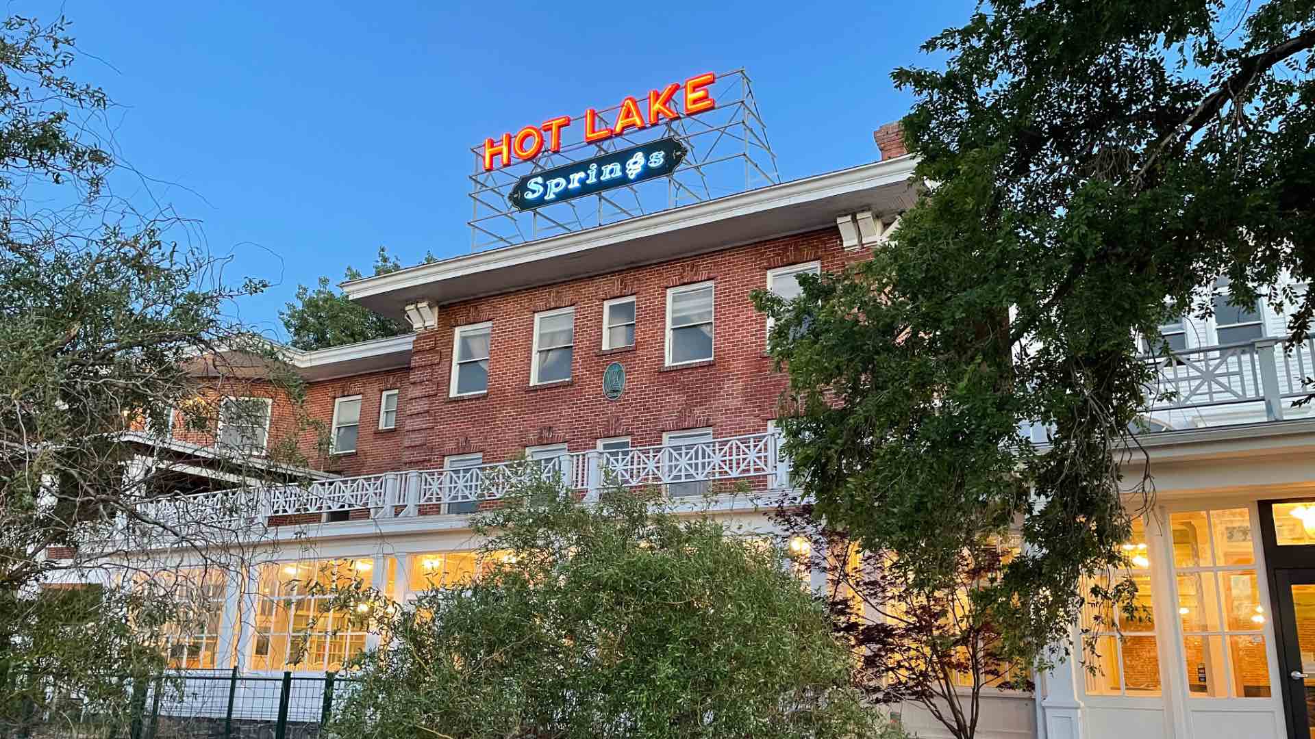 Daytime front view of Hot Lake Springs Resort with neon sign on top, red brick building, and green trees and bushes in foreground.