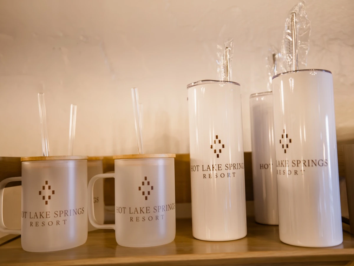Frosted white glass with handle, straw, and Hot Lake Springs Resort logo, next to white thermal bottles with logo on wood shelf.