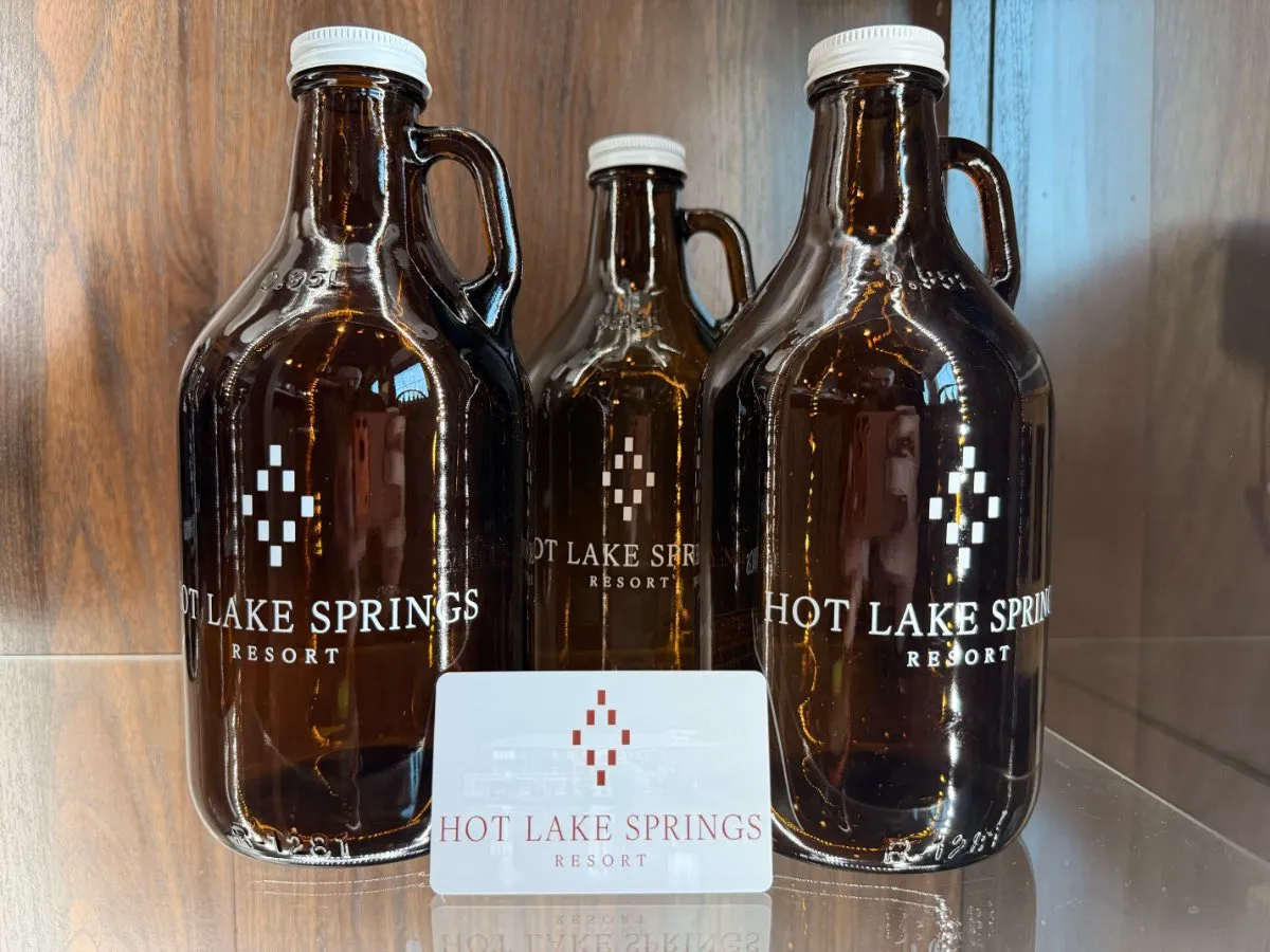 Three dark brown beer growlers with white lids and Hot Lake Springs Resort logo, physical gift card leaning in front.