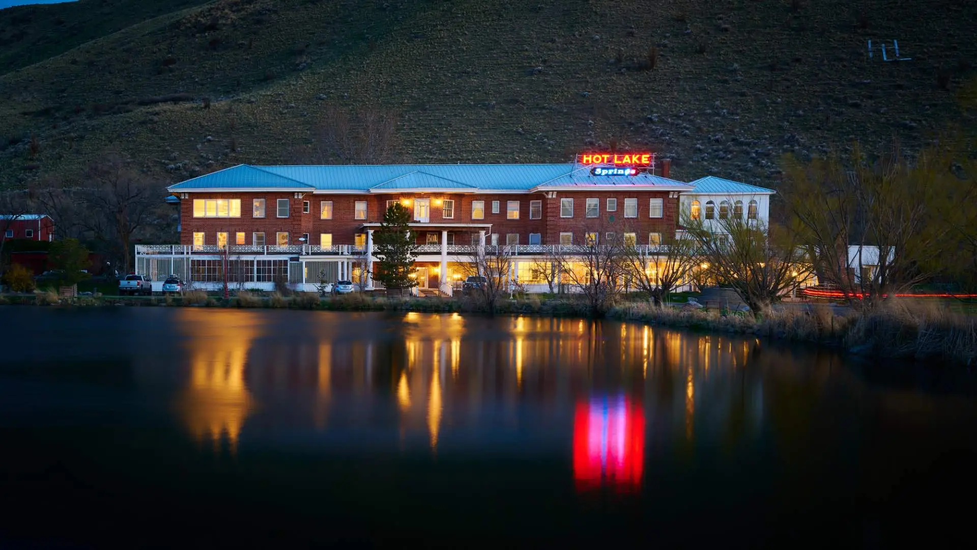 Front exterior of Hot Lake Springs Resort at dusk, red brick facade with iconic neon sign lit up and soft reflection in Hot Lake.
