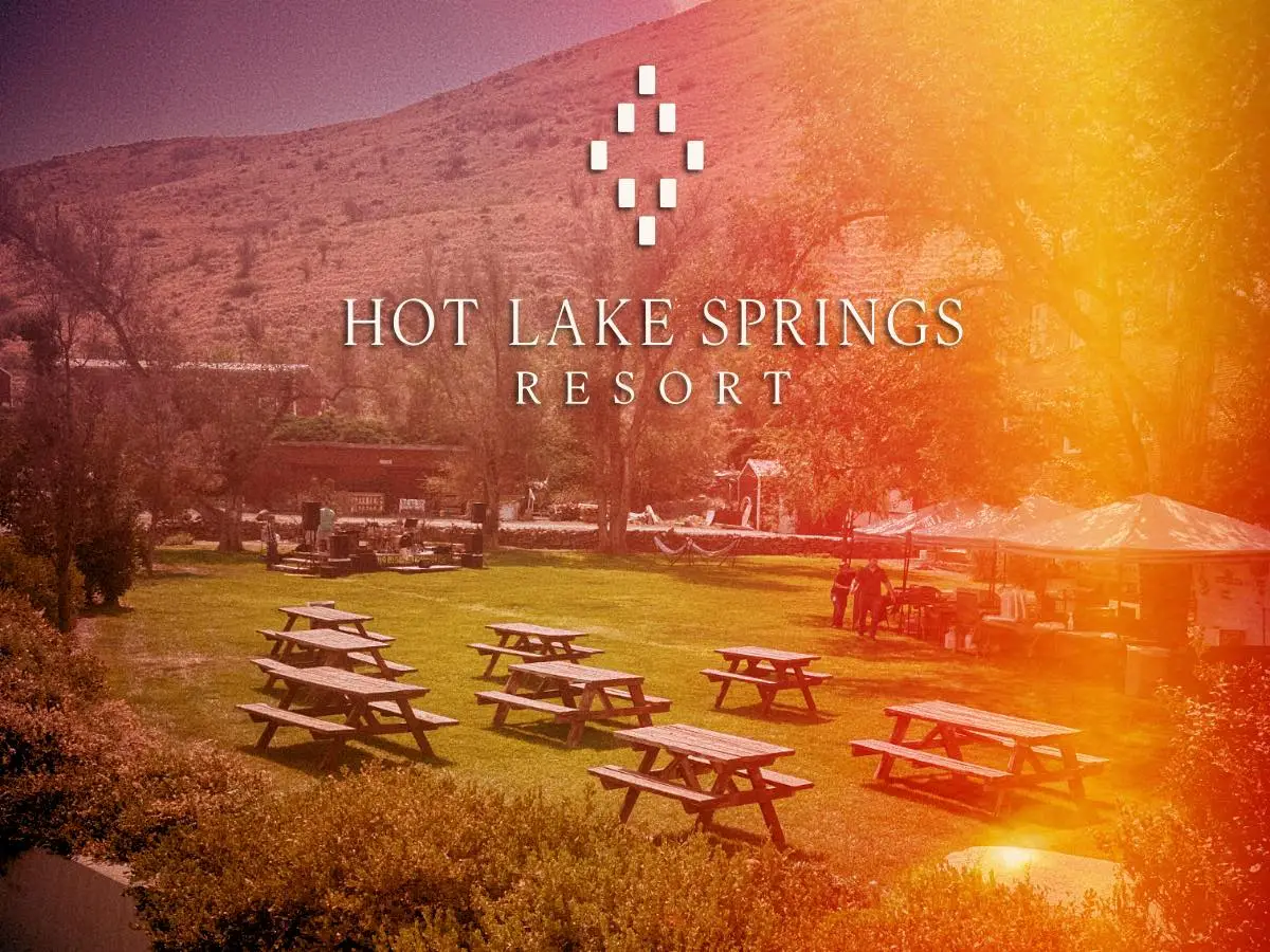 East Lawn at Hot Lake Springs Resort, sunny afternoon, picnic tables on the grass, a stage backed by hillside and trees, orange and yellow light leak across the right side of the frame.